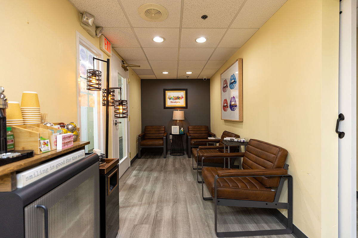 Modern dental office waiting room featuring brown leather chairs, framed artwork, and a coffee and refreshment station.