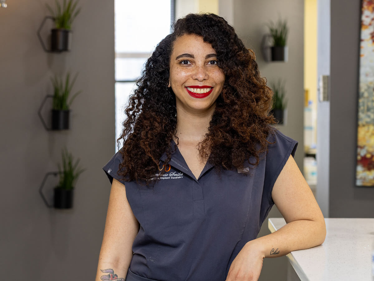 Destiny smiling and leaning against a counter in the Rittenhouse Smiles dental office, wearing short-sleeved dark grey scrubs.