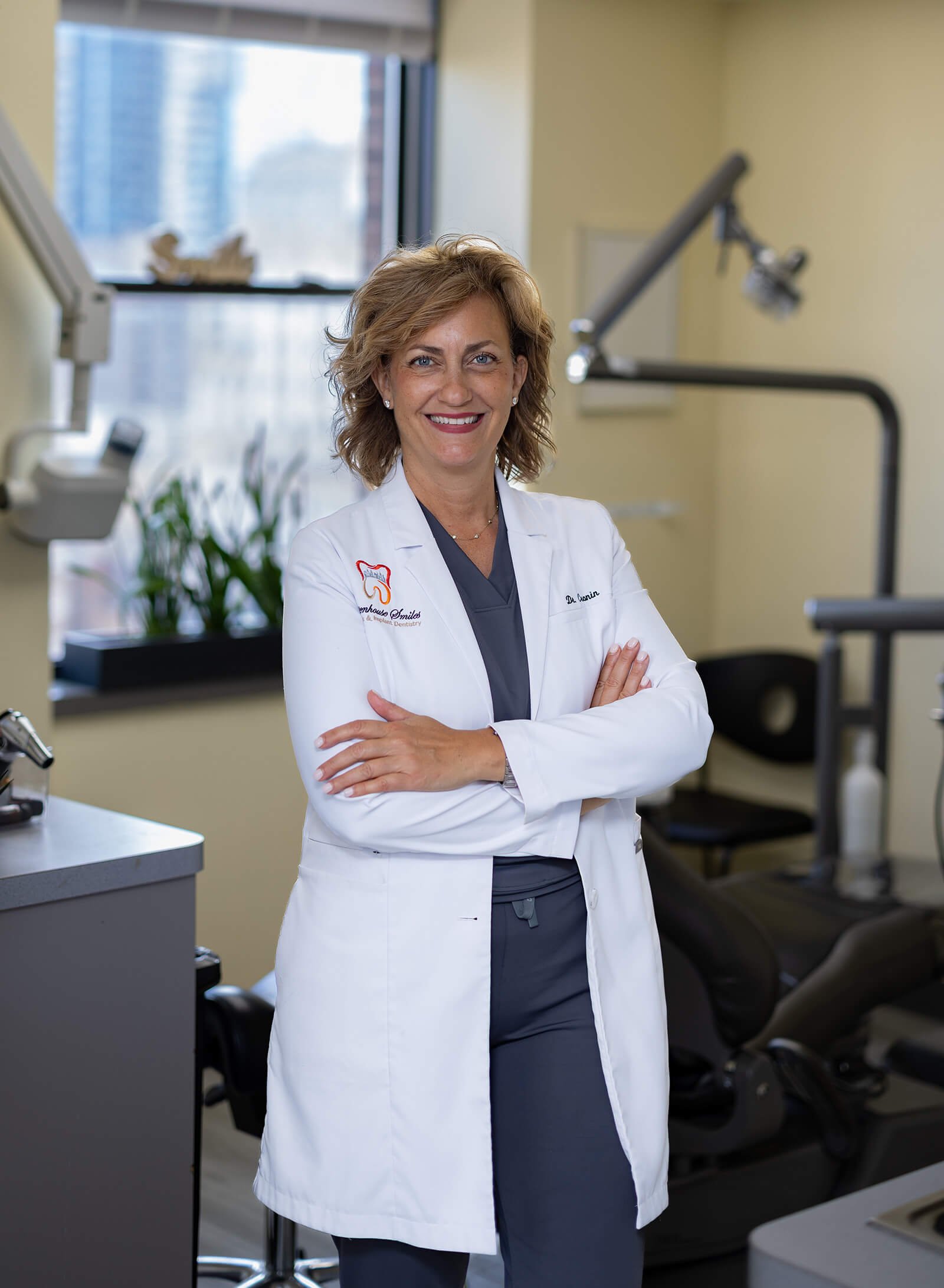 Dr. Andrea Cronin smiling with her arms crossed in a dental treatment room, wearing a white lab coat over dark scrubs.