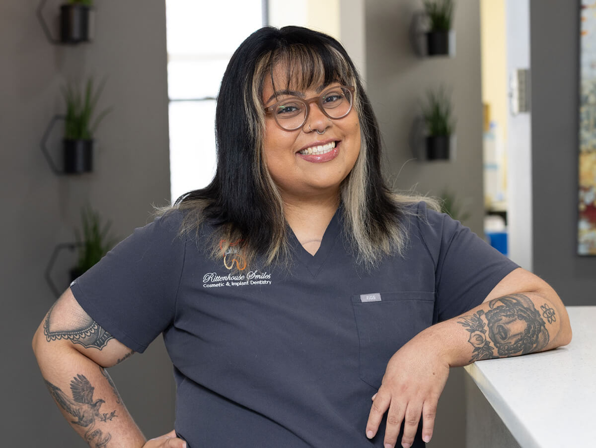 Kat smiling and leaning against a counter in the Rittenhouse Smiles dental office, wearing glasses and short-sleeved dark grey scrubs.
