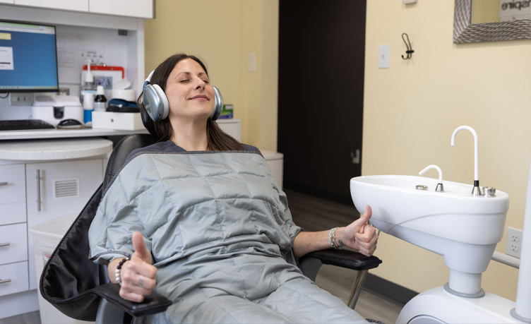 Patient relaxing in a dental chair wearing noise-canceling headphones and a weighted blanket giving two thumbs up