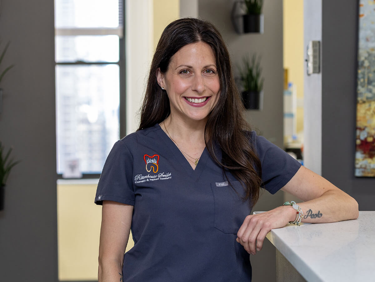 Renee smiling and leaning against a counter in the Rittenhouse Smiles dental office, wearing short-sleeved dark grey scrubs.
