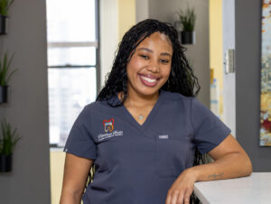 Bri smiling while leaning against a reception counter in a dark gray scrub top embroidered with the Rittenhouse Smiles logo.