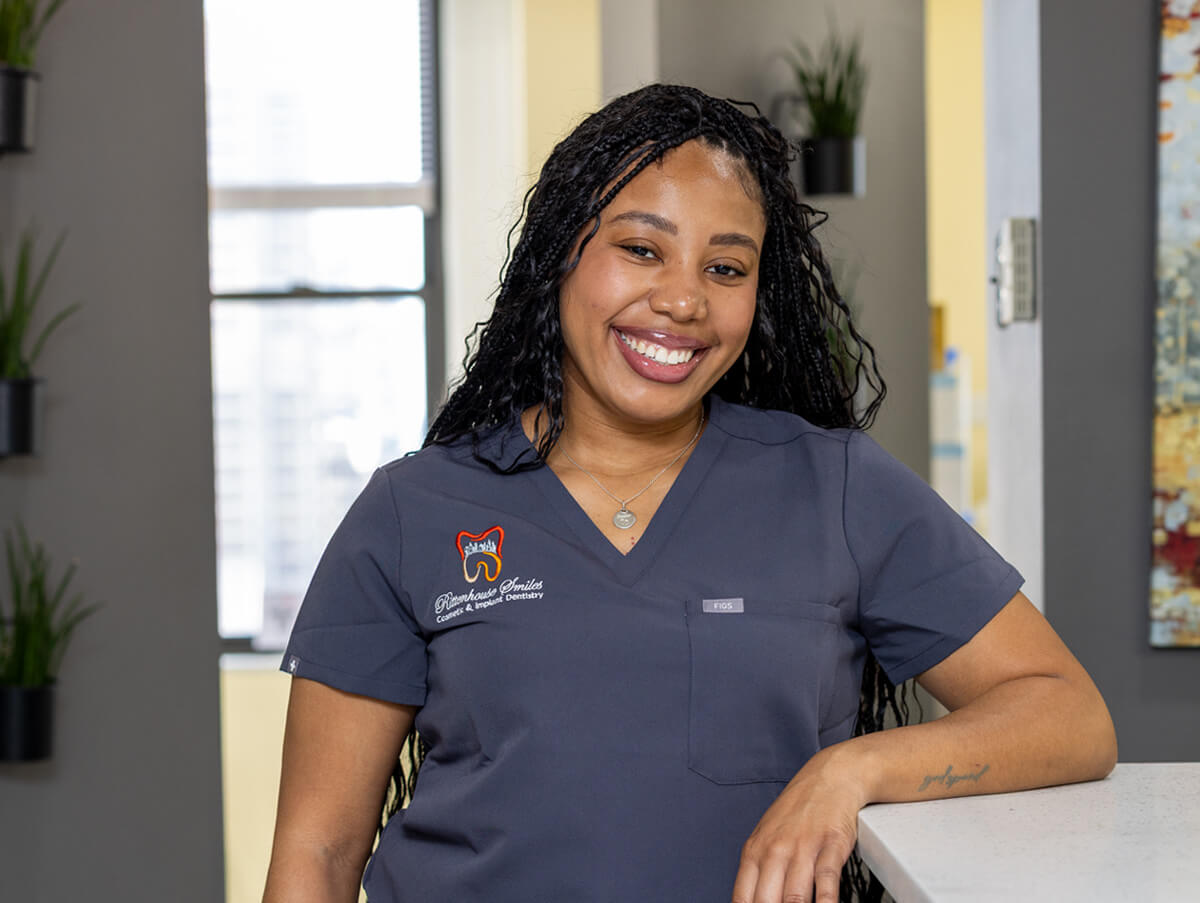 Bri smiling while leaning against a reception counter in a dark gray scrub top embroidered with the Rittenhouse Smiles logo.