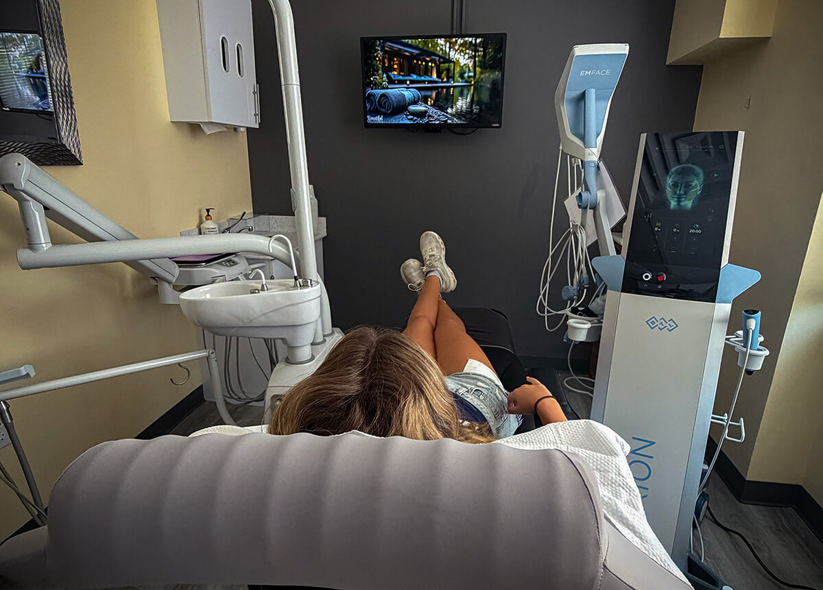 Patient reclining in a treatment chair facing specialized EXION and EMFACE functional wellness technology machines in a modern treatment room.