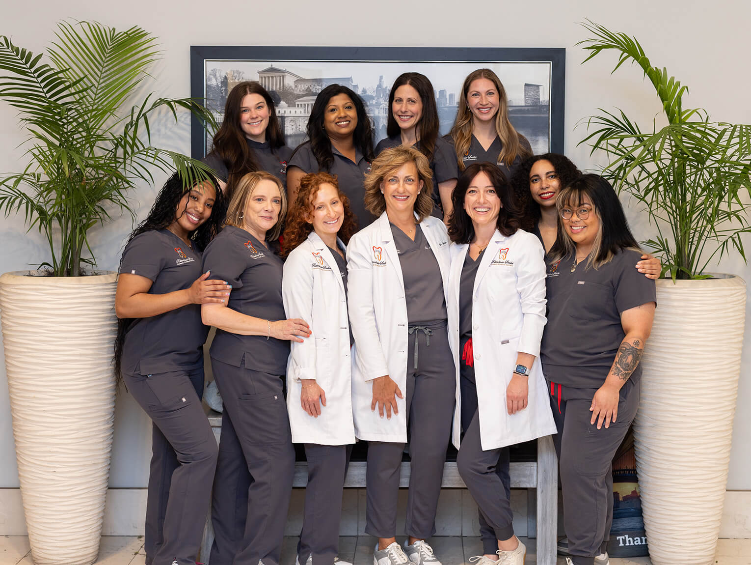 The Rittenhouse Smiles dental team smiling together in the lobby, featuring three doctors in white lab coats and seven team members in dark grey scrubs.