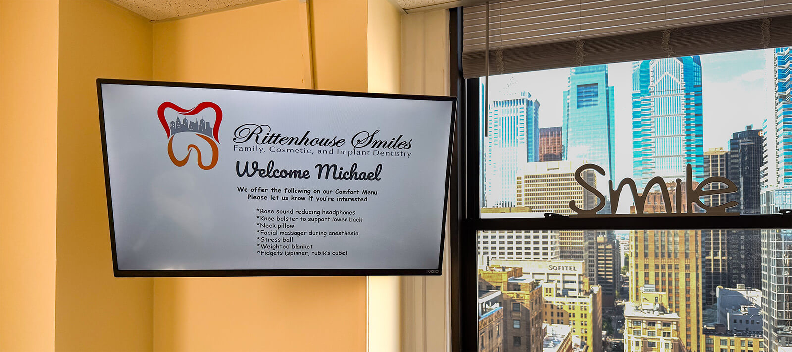 A wall-mounted TV screen in a treatment room displaying a personalized "Welcome Michael" message alongside a Rittenhouse Smiles Comfort Menu, which lists patient amenities such as noise-reducing headphones, a neck pillow, and a calming lap dog.